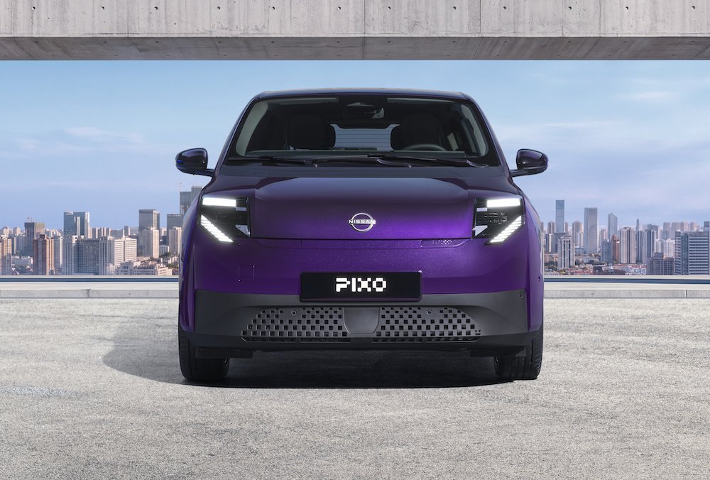 Photograph showcasing a purple Nissan Pixo concept car parked before a concrete architectural opening with a city skyline beyond. Front view highlights illuminated angular headlights, closed grille, PIXO plate, sleek compact design, and blue sky backdrop.